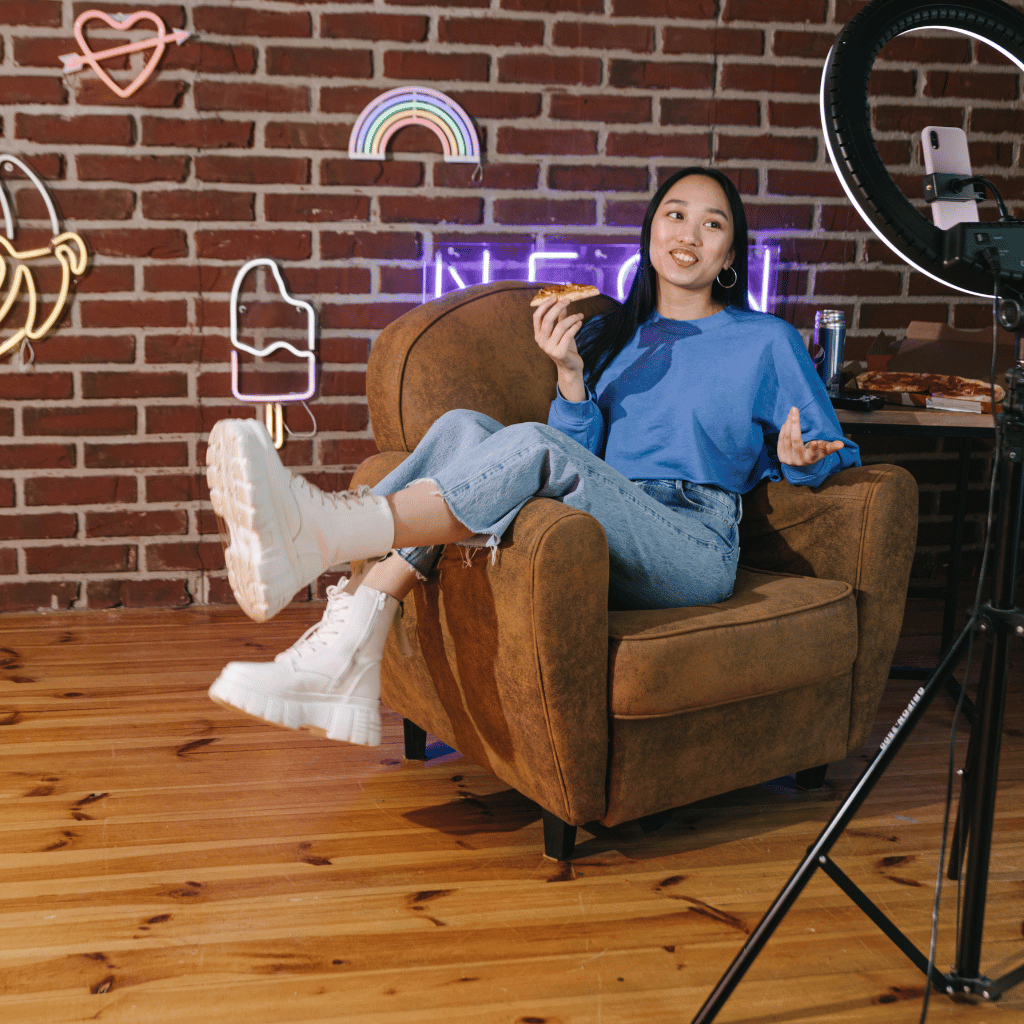 A young woman in casual attire lounges in an armchair, animatedly speaking to a smartphone on a tripod with a ring light, in a cozy room adorned with neon signs.