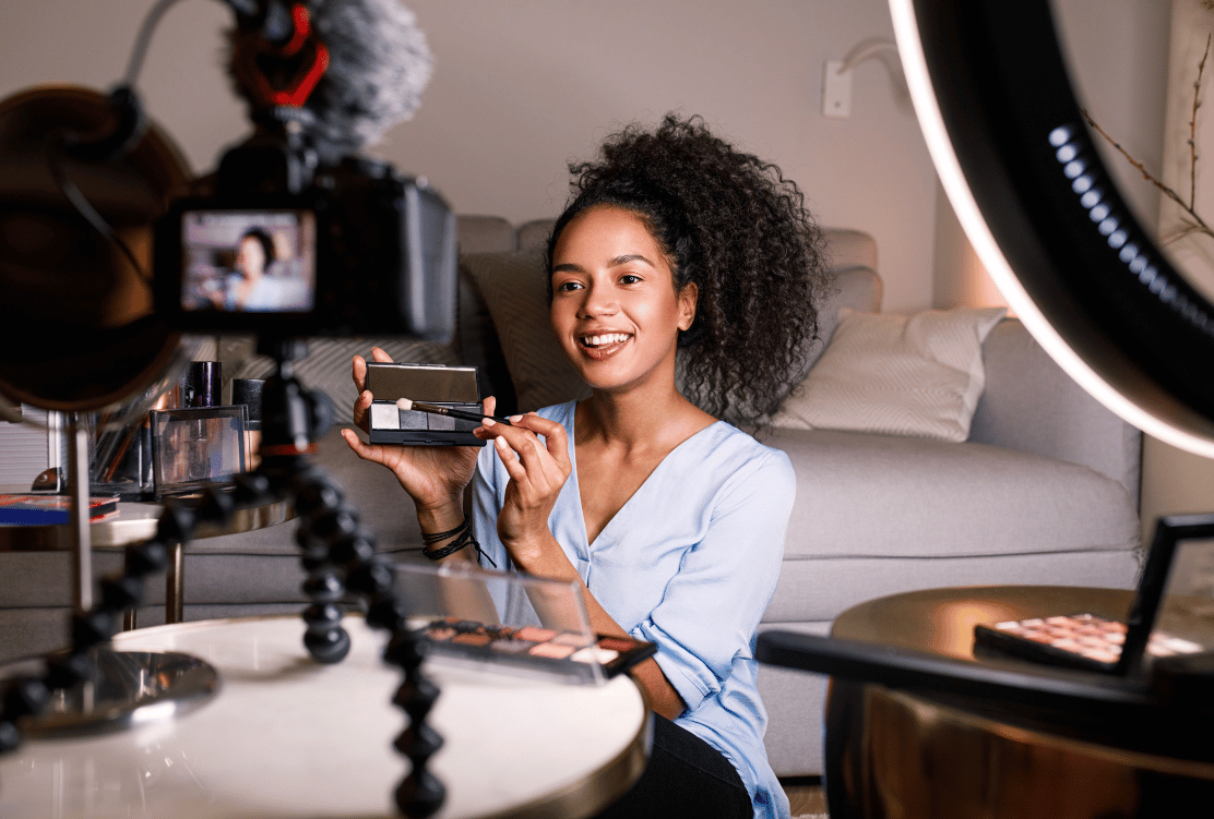 A smiling woman showcasing makeup products while recording a video with a DSLR camera, set up on a tripod with a ring light.