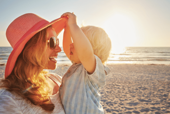 A smiling woman wearing sunglasses and a sunhat holds a young child at the beach, both basking in the golden glow of the sunset.