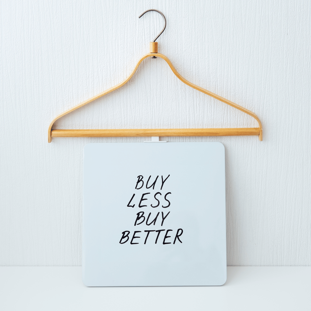 A wooden hanger holds a square sign with the minimalist message 'BUY LESS BUY BETTER' against a white textured background.