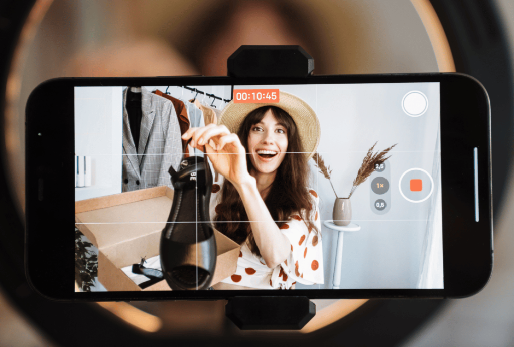 Woman in a polka-dot dress and hat smiling and recording a fashion vlog on a smartphone, with a focus on stylish footwear.