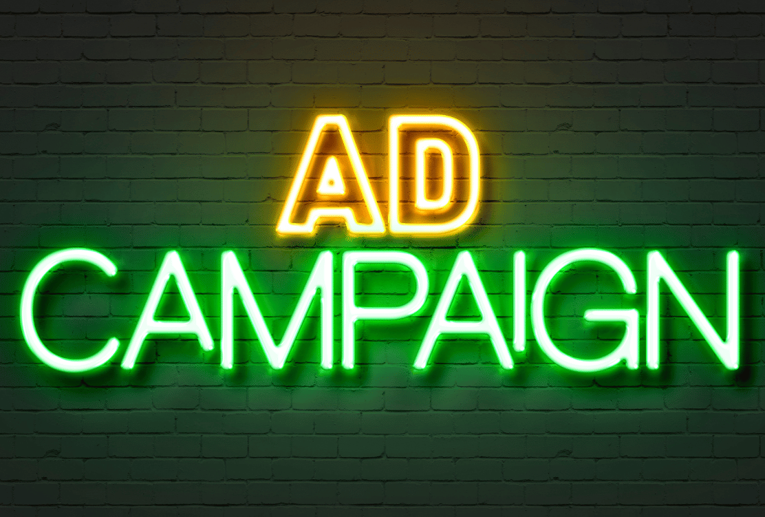 Neon sign on a brick wall displaying the words "AD CAMPAIGN" in bright yellow and green lights.