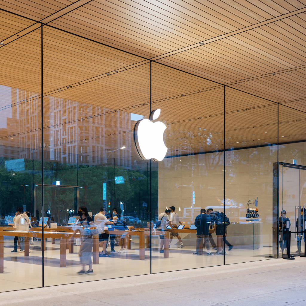 Customers engage with products in a brightly lit Apple Store, visible through its iconic glass facade featuring a large Apple logo.