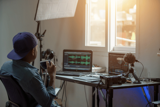 A content creator sits at a desk facing a laptop with audio editing software on screen, surrounded by podcasting equipment, in a home studio setup.