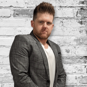 A confident man standing against a white brick wall background.