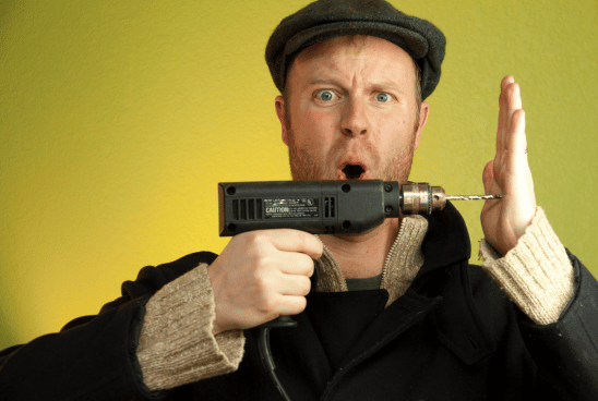 A startled man wearing a flat cap holds a power drill close to his ear, symbolizing a poor decision.