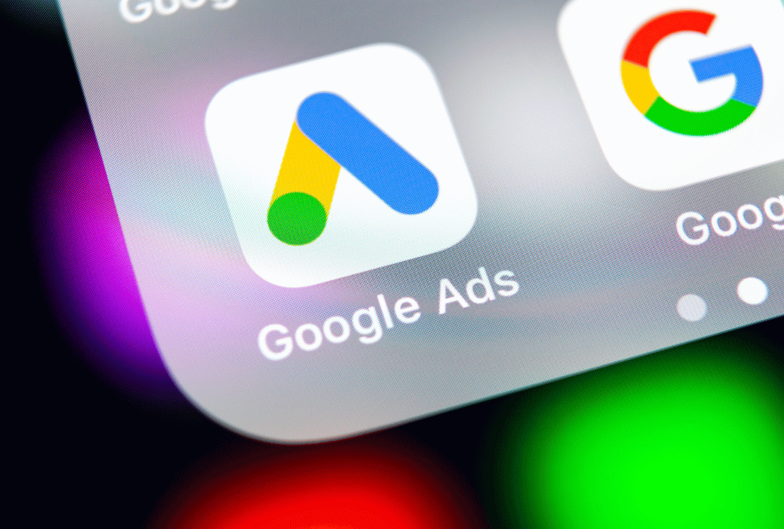 Close-up of the Google Ads app icon on a smartphone screen, with other Google service icons blurred in the background.