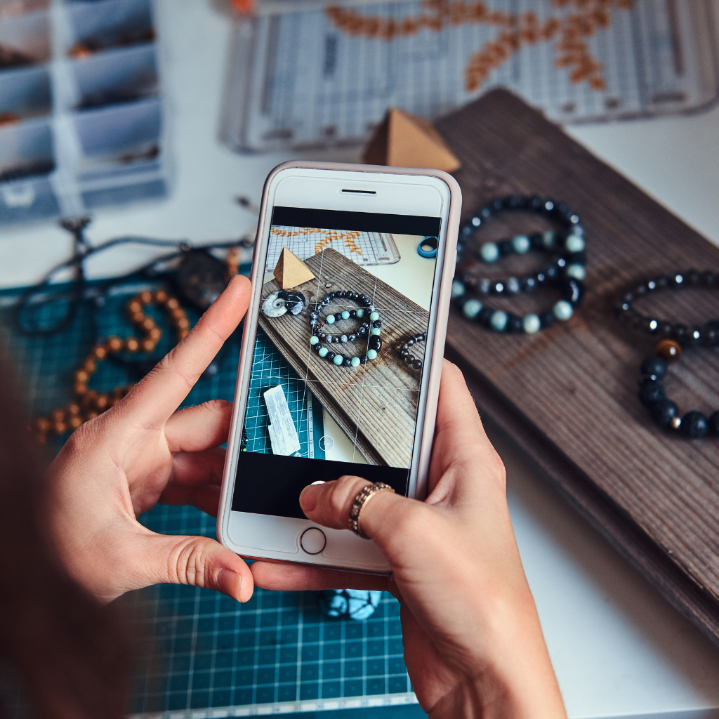 Close-up of hands using a smartphone to take a photo of handmade jewelry on a crafting table.