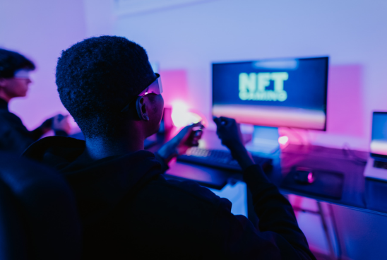 A person in profile view, wearing glasses and a hoodie, is focused on a computer screen with the words "NET Gaming" displayed, indicative of a gaming environment or an esports event.