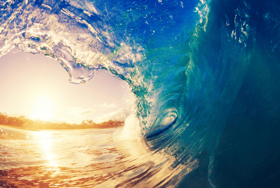 The curl of a crystal clear blue wave captured from inside, with the sun setting in the background.