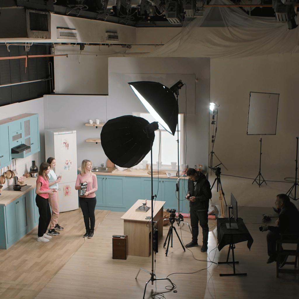 Behind-the-scenes view of a kitchen set for a video production with lighting equipment, camera crew, and talent in preparation.