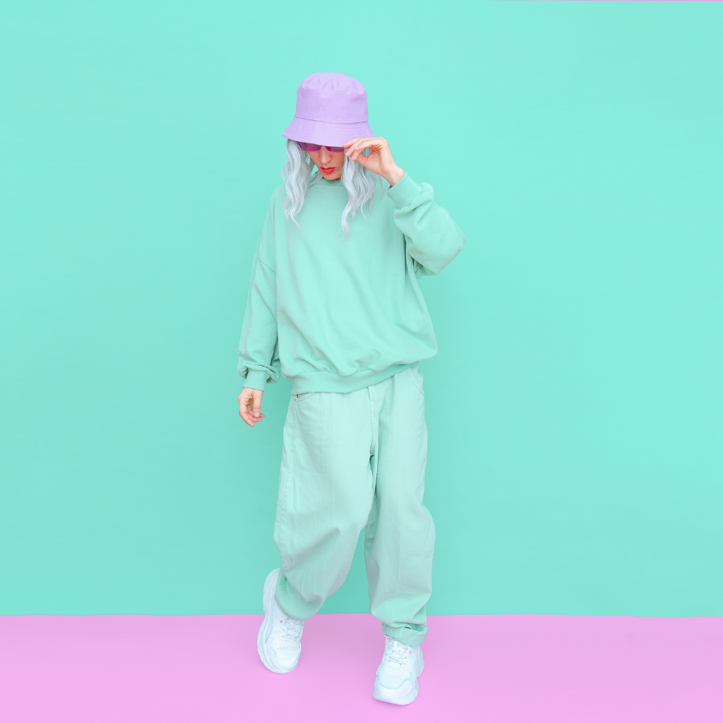 A fashion-forward individual in pastel streetwear poses, with their hand adjusting a lilac bucket hat, set against a turquoise and pink background.