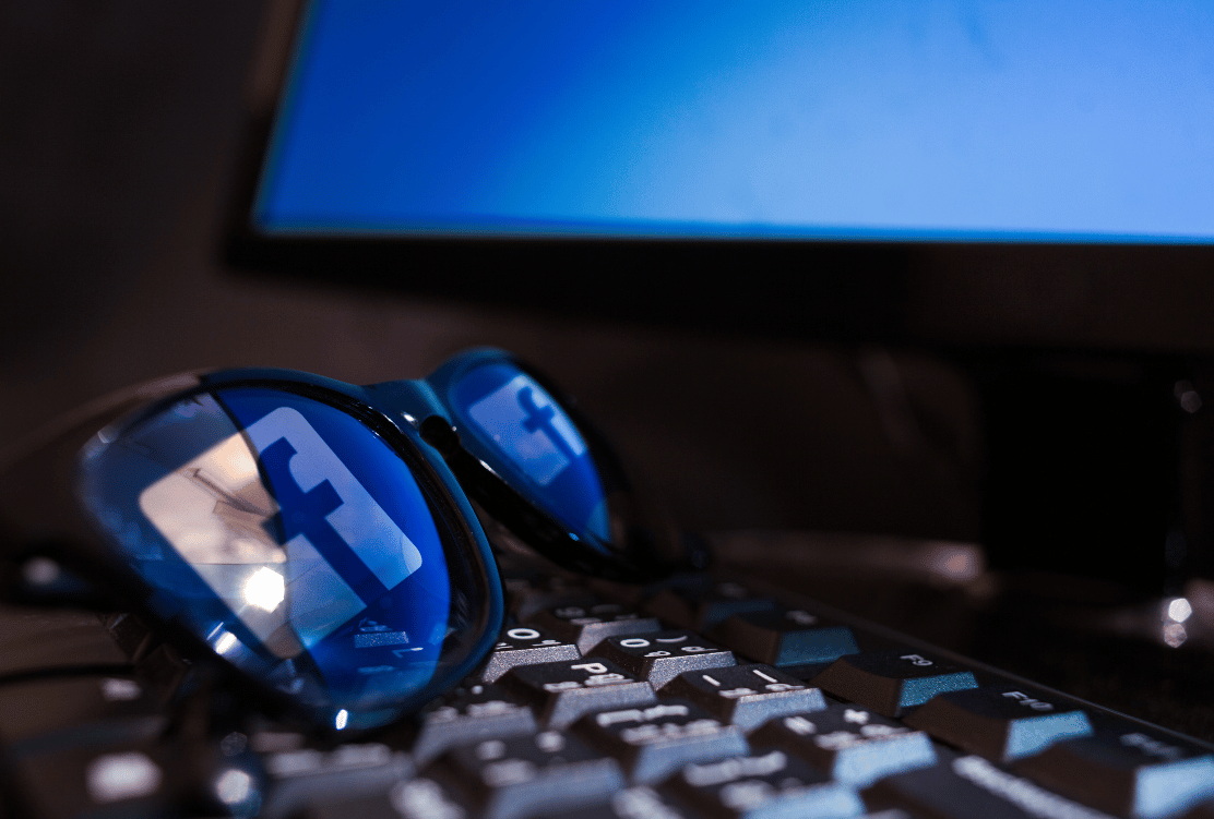 Prescription glasses with Facebook logos reflecting on the lenses, resting on a backlit keyboard with a computer monitor in the background.