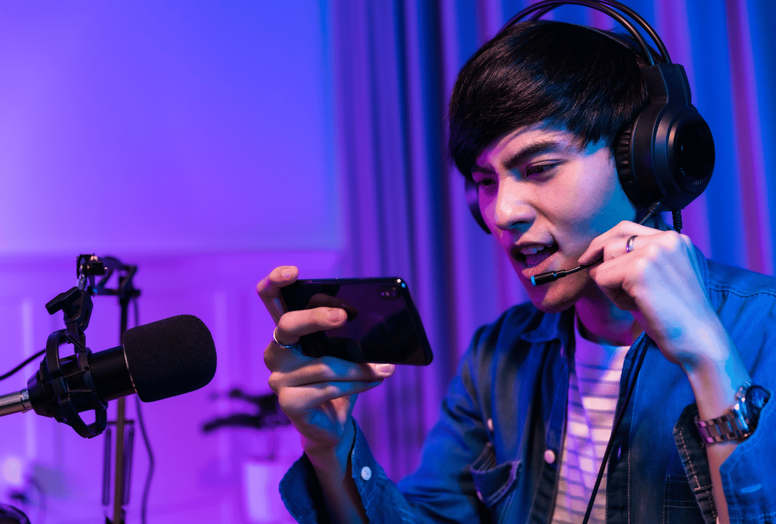 A focused young man with headphones and a microphone creating content on his smartphone, in a studio illuminated with purple lighting.