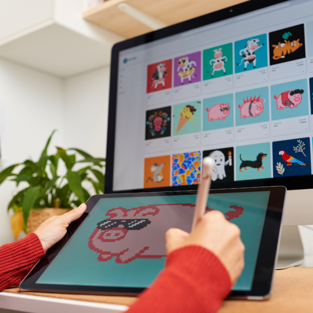 An artist creates a pixel art design on a tablet, with a monitor in the background displaying a selection of colorful NFT artworks.