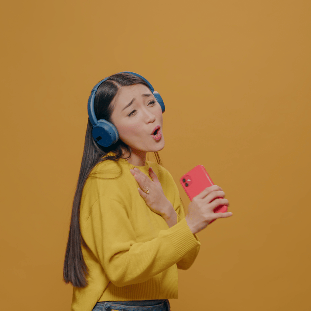 A young woman in a yellow sweater and blue headphones looks animatedly at her pink smartphone, appearing to sing or speak passionately.