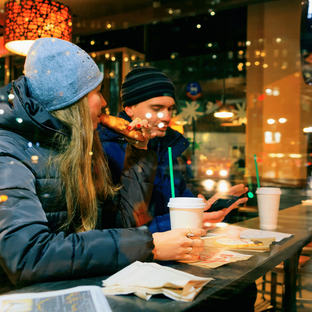 Two people in winter attire enjoy food and drinks at a Starbucks, with one person browsing their phone, highlighting a relaxed, customer-friendly environment.