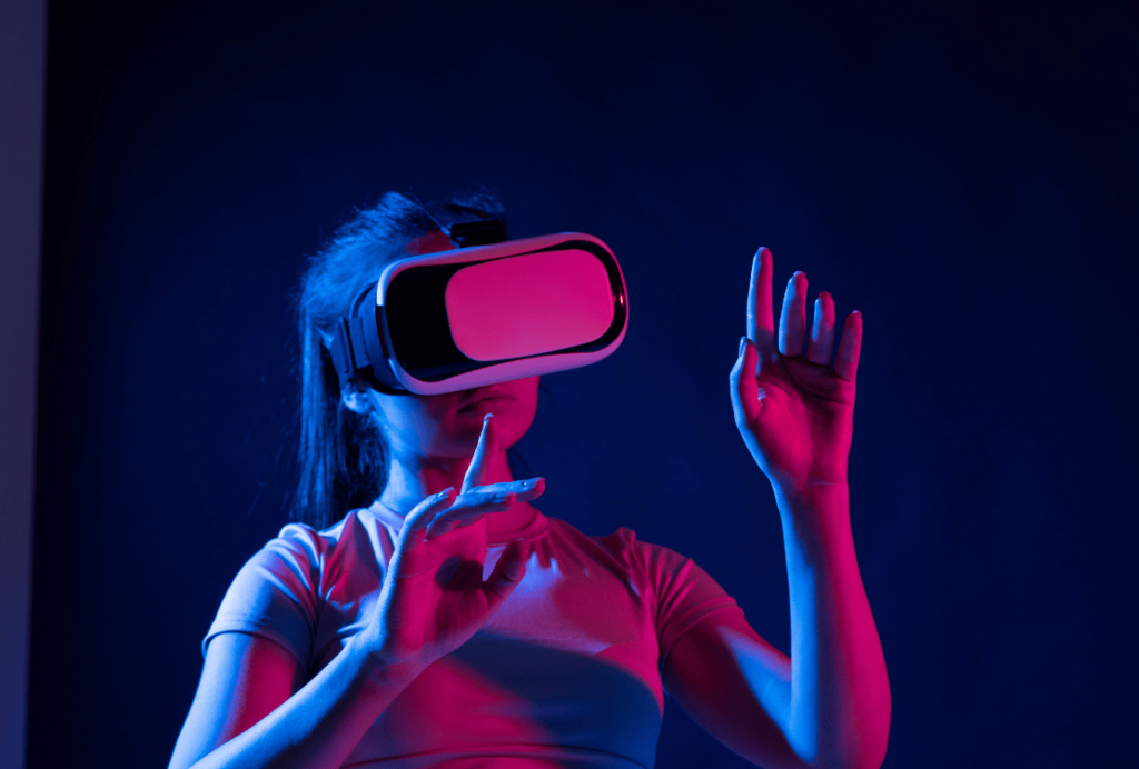 A woman in a white t-shirt engages with virtual reality, gesturing with her hands, against a dark background illuminated by neon lights.