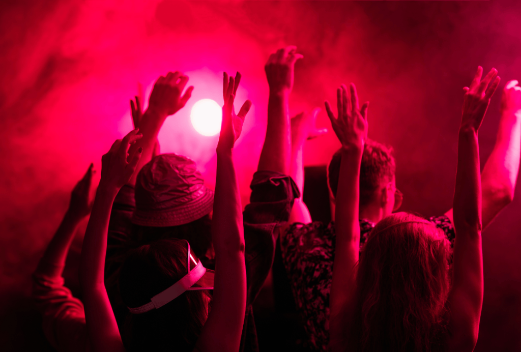 A crowd of people at a concert raising their hands towards a glowing light with a red haze.