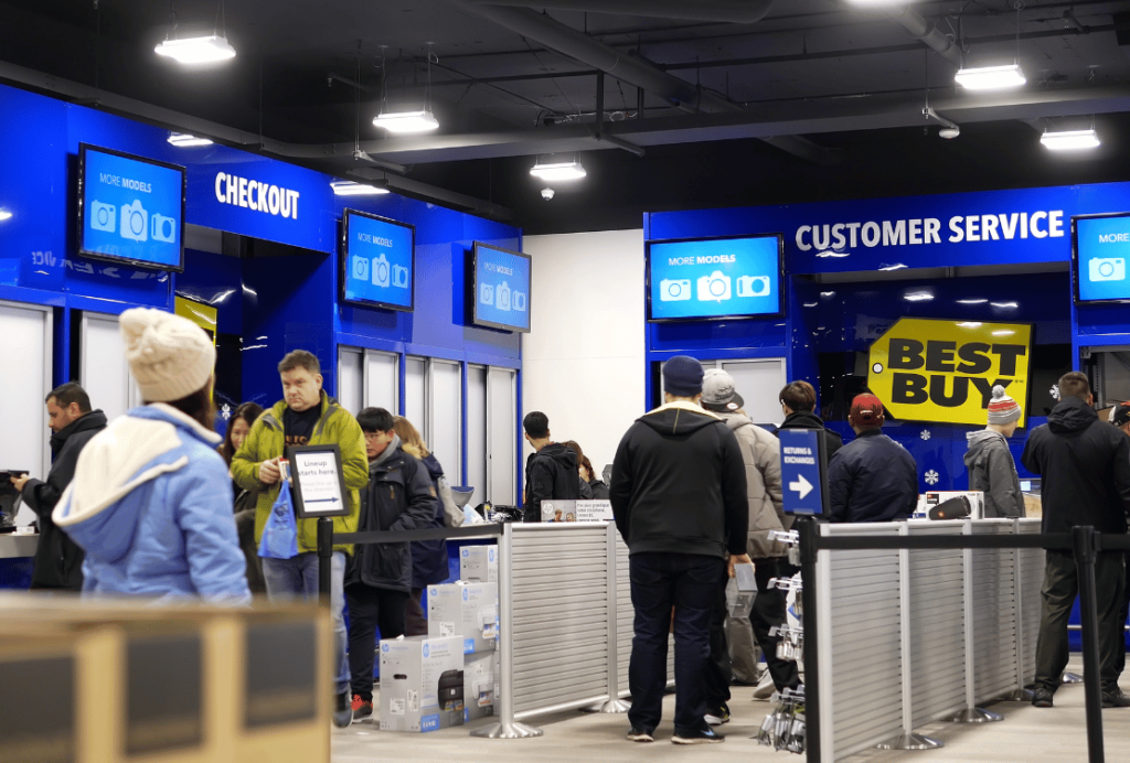 Customers lined up at a Best Buy checkout counter with blue signage indicating customer service and more models available.