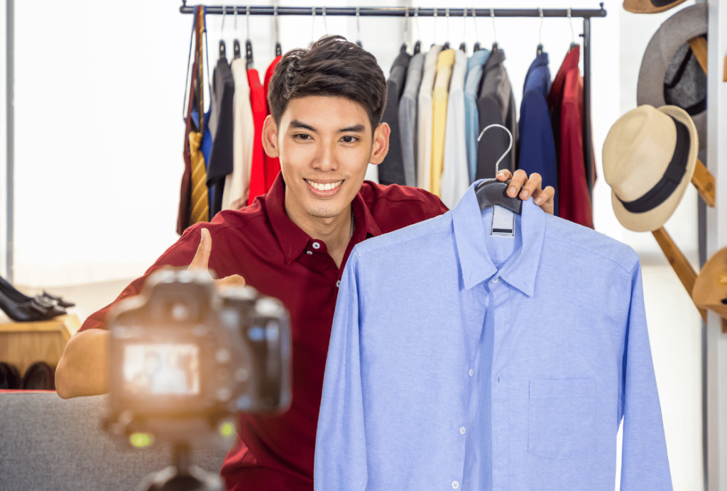 A smiling man presenting a blue shirt to a camera with a rack of clothes in the background.