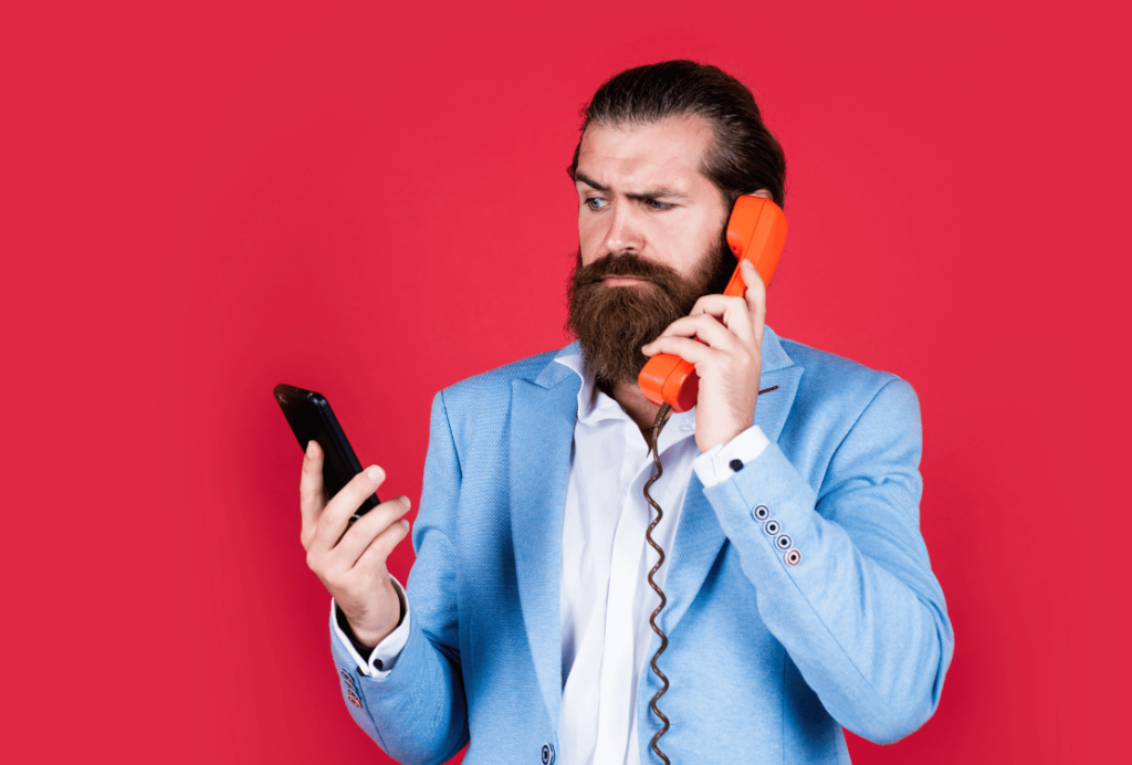 A confused bearded man in a blue suit holding a traditional orange handset to his ear and a smartphone in the other hand against a red background.