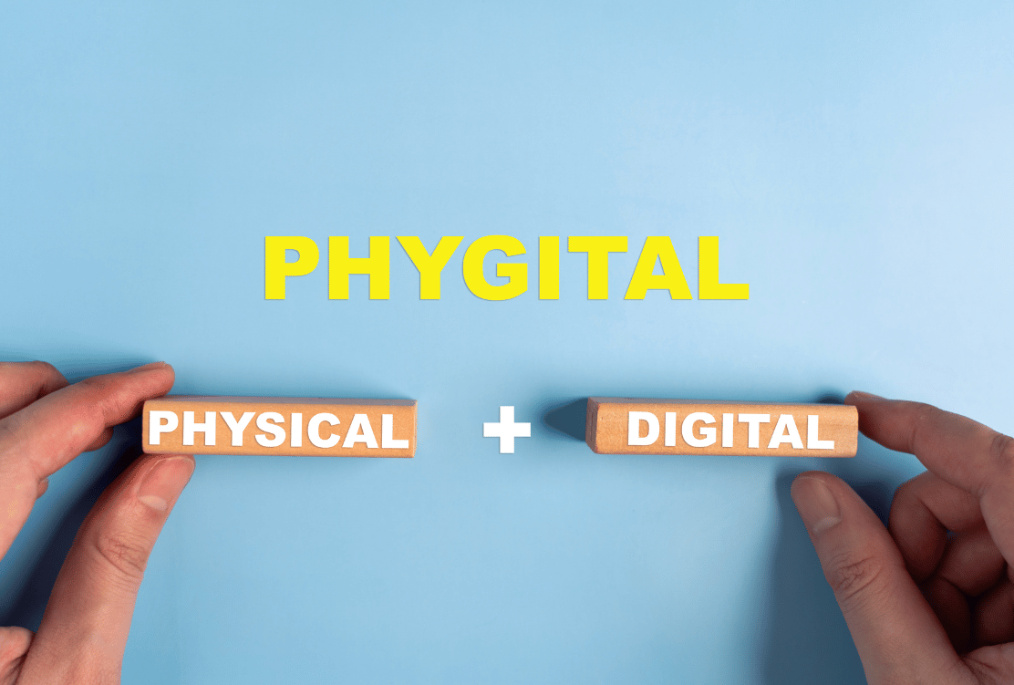 Two hands holding wooden blocks that spell out 'PHYSICAL' and 'DIGITAL', combining to form the word 'PHYGITAL' displayed above in bold yellow letters on a blue background.