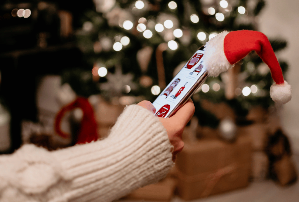 A person browsing holiday deals on a smartphone with a Santa hat perched on top, in front of a blurred Christmas tree background.