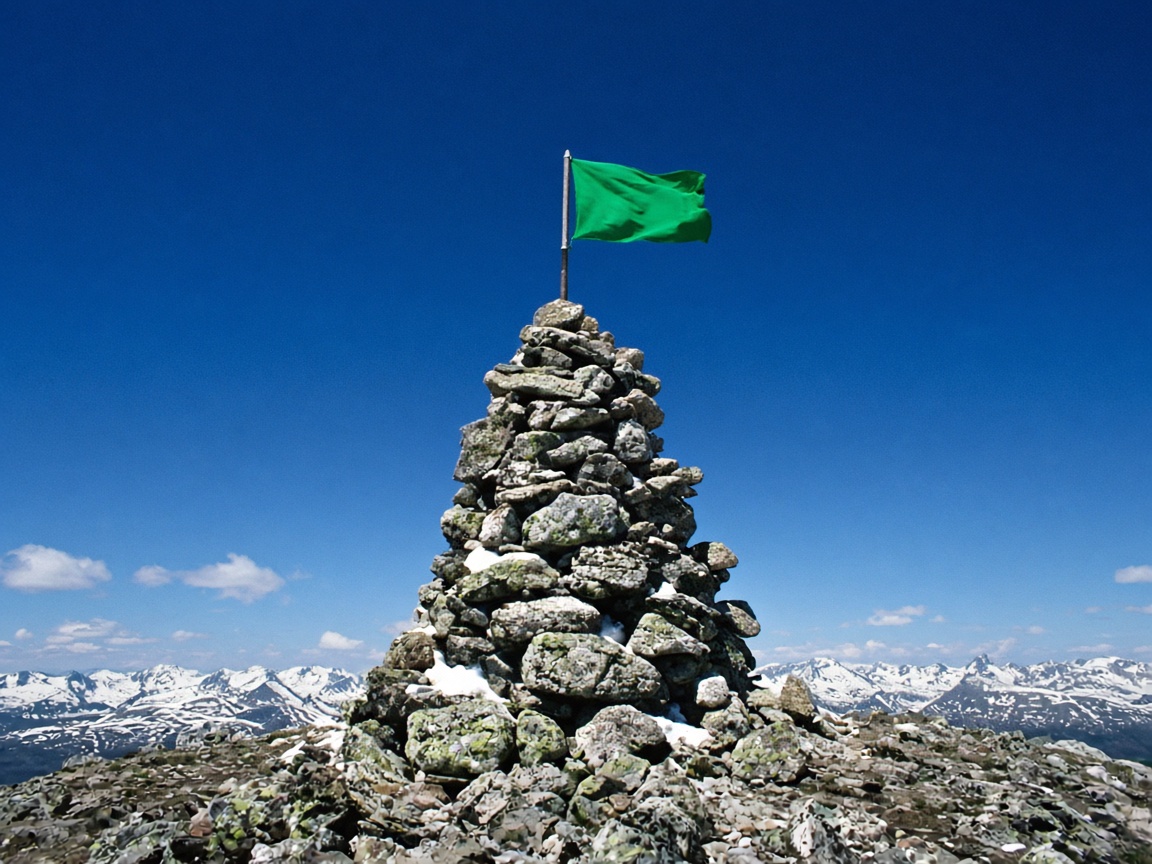 A tall summit cairn of stacked grey stones on a mountain ridge with a single new emerald flag planted on top, snapping in the wind under a deep blue sky.