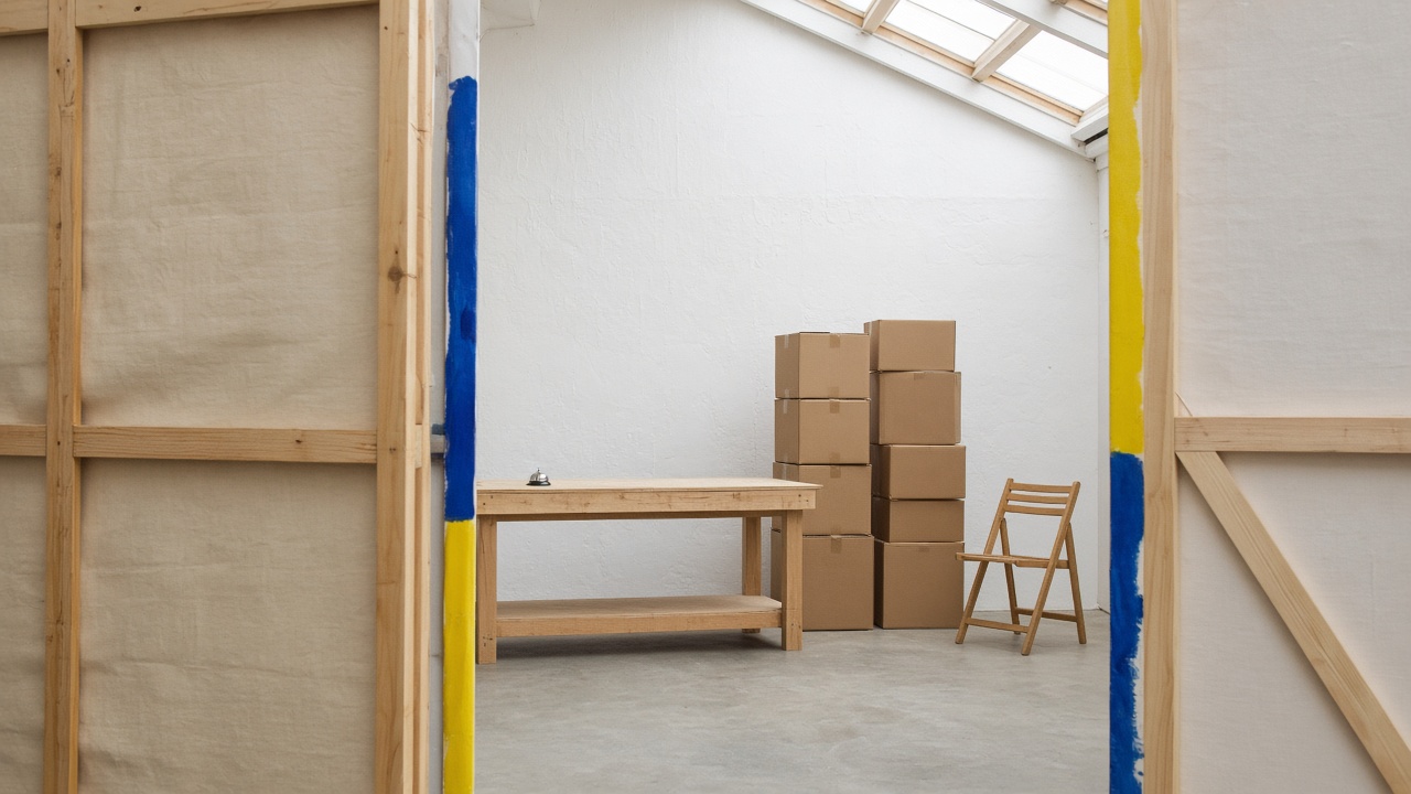The back of a stage set in daylight, with timber braces, plain unmarked parcel boxes, a folding chair and a small service bell on a bare counter.